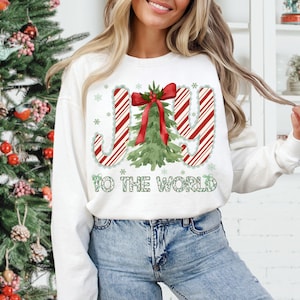 Puede incluir: Sudadera blanca con un gr&aacute;fico navide&ntilde;o. El dise&ntilde;o presenta la palabra "JOY" en rayas rojas y blancas de bast&oacute;n de caramelo, un &aacute;rbol de Navidad verde y la frase "TO THE WORLD" en verde. Los acentos de copo de nieve completan el look festivo.