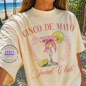 May include: Cream-colored t-shirt with a Cinco de Mayo design. The graphic features a margarita with a pink bow, lime, and the words "Cinco de Mayo Social Club" in pink script. The shirt has a grid pattern.