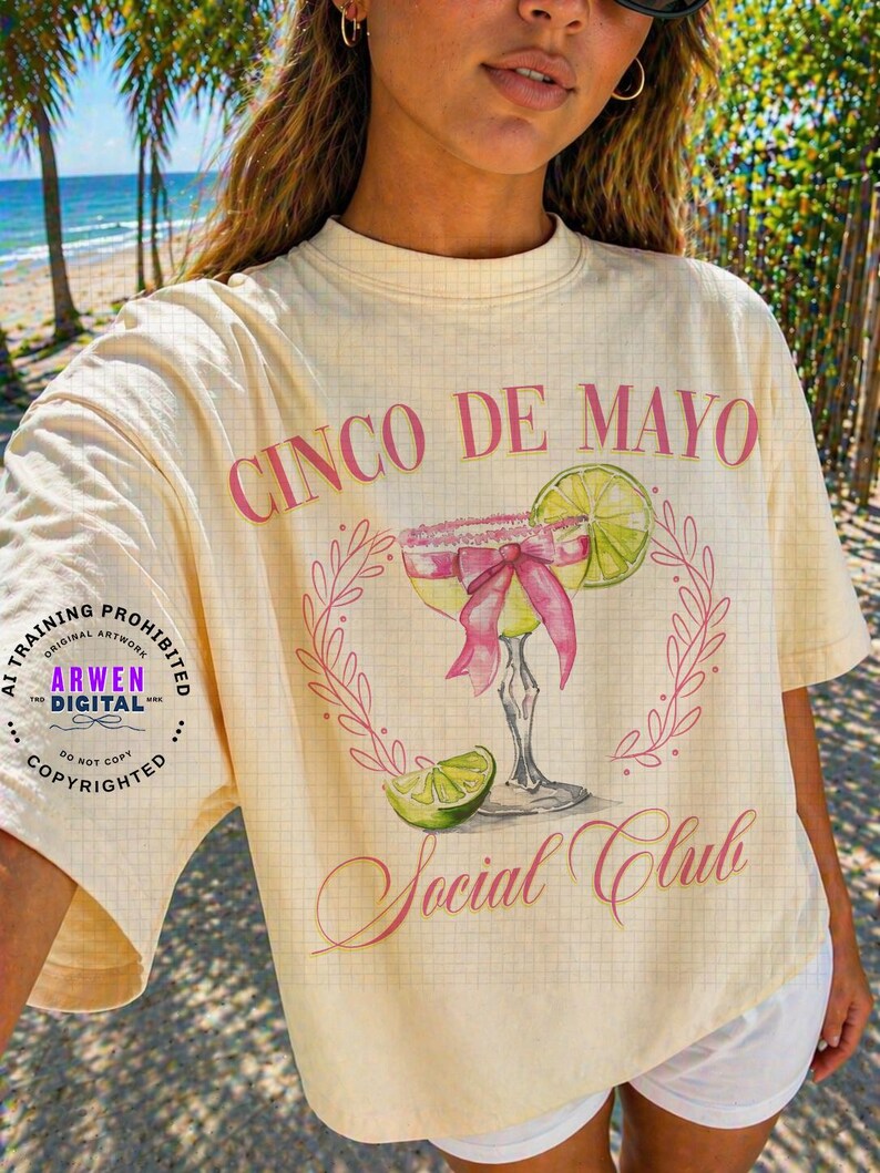 May include: Cream-colored t-shirt with a Cinco de Mayo design. The graphic features a margarita with a pink bow, lime, and the words "Cinco de Mayo Social Club" in pink script. The shirt has a grid pattern.