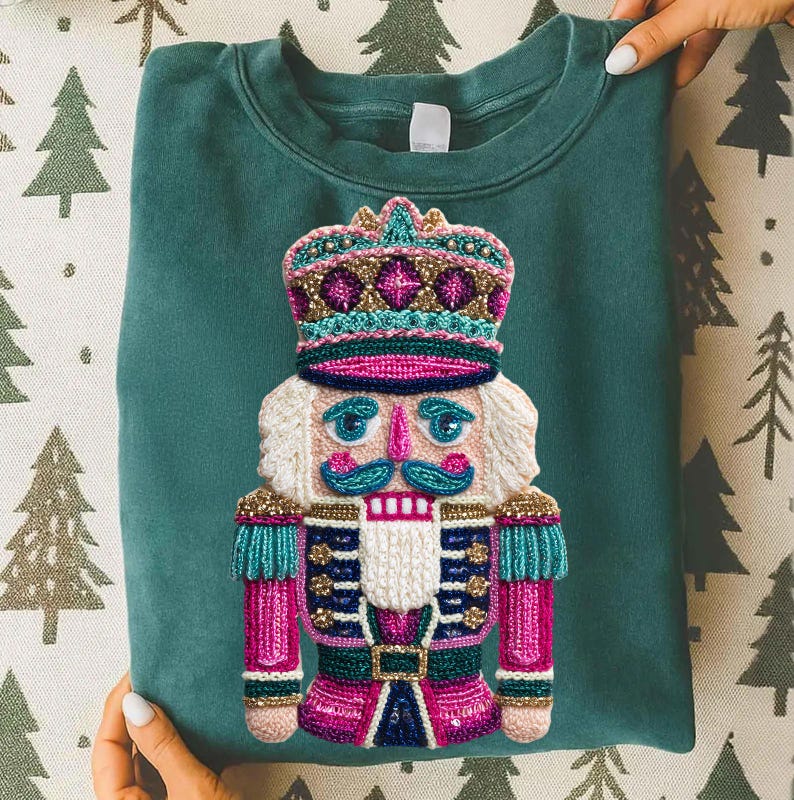 May include: Teal green sweatshirt featuring a colorful, detailed embroidered nutcracker design. The nutcracker has a crown, mustache, and uniform in shades of pink, teal, gold, and white. The sweatshirt is held by hands with white nail polish.