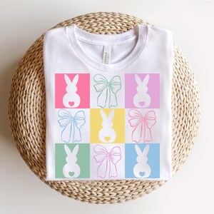 May include: White t-shirt with a colorful design of bunnies and bows. The bunnies are pink, green, blue, and yellow. The bows are pink, blue, green, and yellow.
