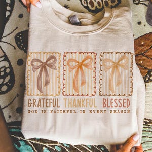 May include: A beige sweatshirt featuring three square designs with watercolor bows in shades of brown and peach. The text "Grateful Thankful Blessed" and "God is faithful in every season" are printed below the bows.