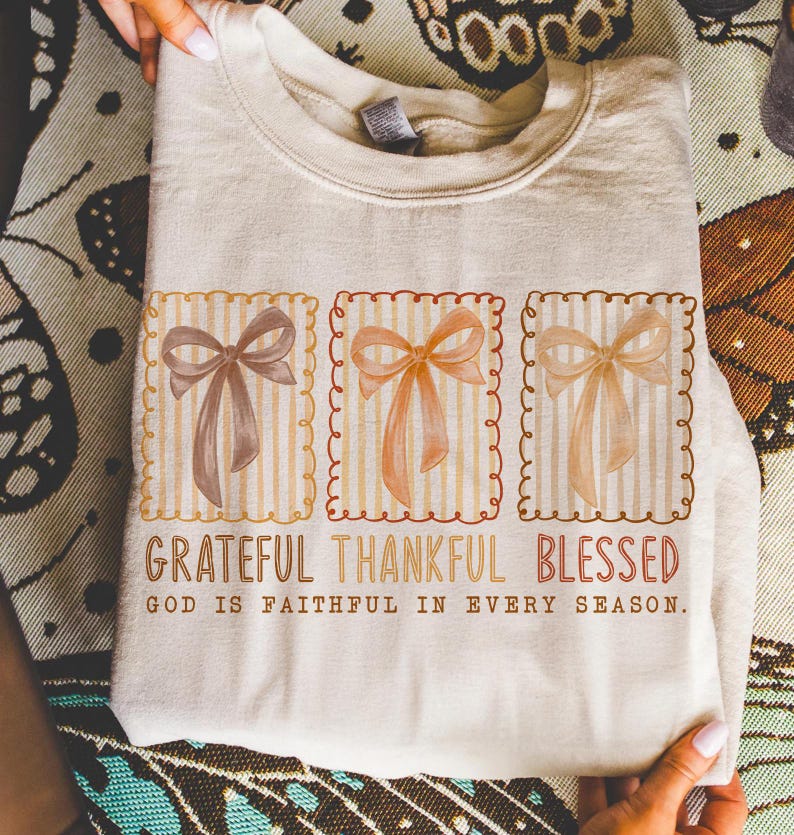 May include: A beige sweatshirt featuring three square designs with watercolor bows in shades of brown and peach. The text "Grateful Thankful Blessed" and "God is faithful in every season" are printed below the bows.