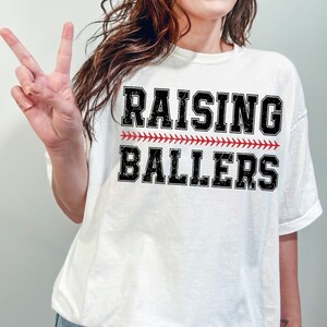 May include: White t-shirt with black text that reads "Raising Ballers" with a red baseball stitch design.