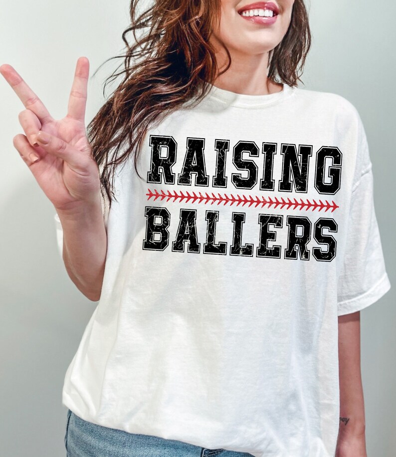May include: White t-shirt with black text that reads "Raising Ballers" with a red baseball stitch design.