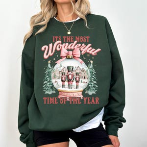 May include: Green Christmas sweatshirt with a pink and white bauble design. The bauble features three nutcracker figures and the text "It's the most Wonderful Time of the Year."