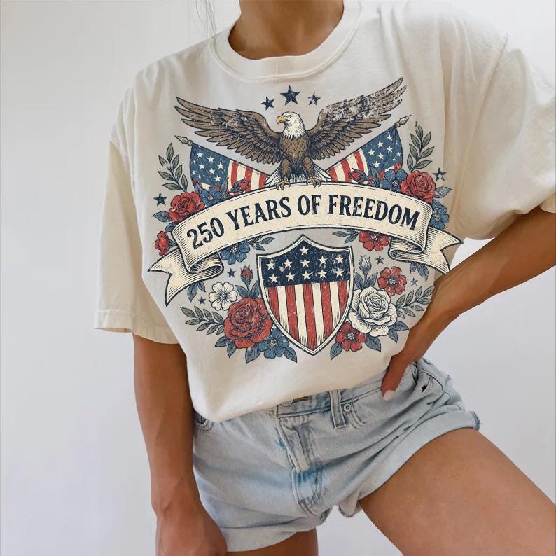 May include: Cream-colored t-shirt featuring a patriotic design. The graphic includes an eagle, American flags, a shield, and the text "250 YEARS OF FREEDOM" on a banner, surrounded by red and blue floral elements.