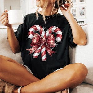 Op de afbeelding: Zwart T-shirt met een feestelijk ontwerp van twee zuurstokken en een grote rode strik met glitteraccenten. De zuurstokken hebben rood-witte strepen. De strik is dieprood met sprankelende details.