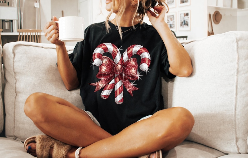 Op de afbeelding: Zwart T-shirt met een feestelijk ontwerp van twee zuurstokken en een grote rode strik met glitteraccenten. De zuurstokken hebben rood-witte strepen. De strik is dieprood met sprankelende details.