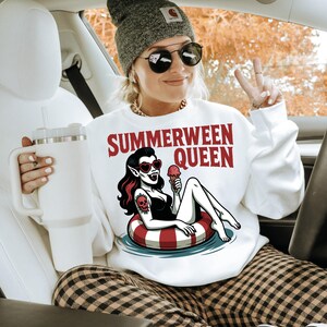 May include: White sweatshirt with the text "SUMMERWEEN QUEEN" and a graphic of a vampire woman in a pool float. The woman is wearing sunglasses and holding an ice cream cone. The person in the image is wearing a gray beanie and sunglasses.