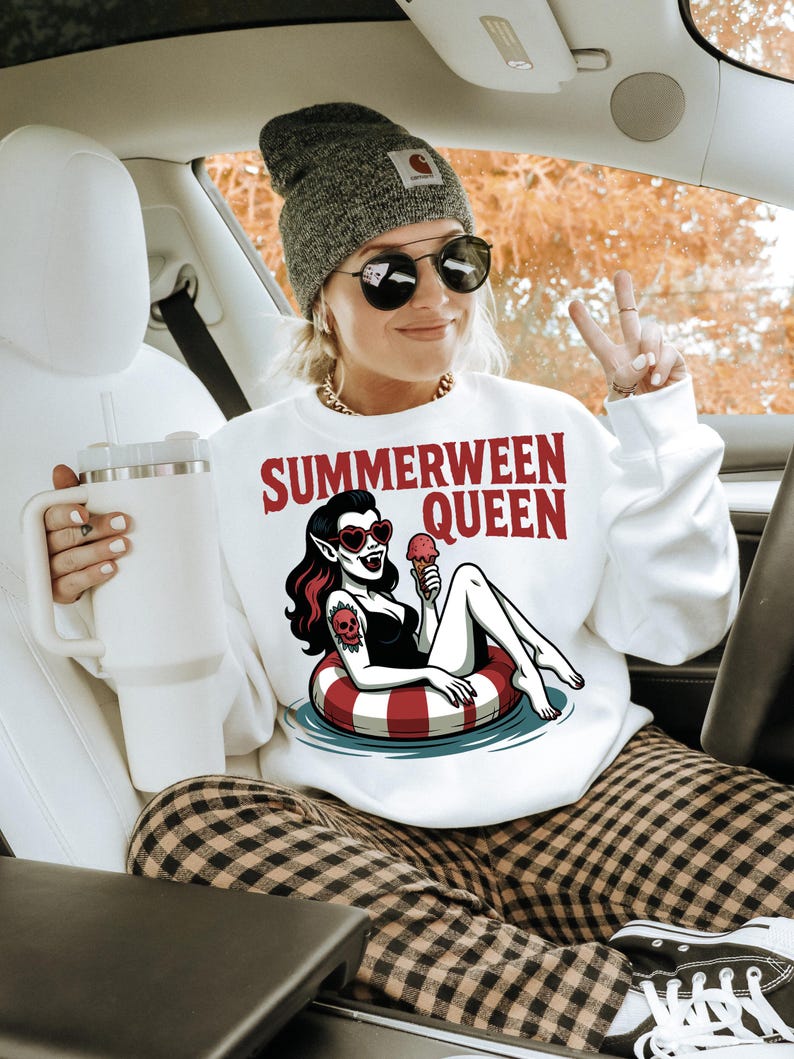 May include: White sweatshirt with the text "SUMMERWEEN QUEEN" and a graphic of a vampire woman in a pool float. The woman is wearing sunglasses and holding an ice cream cone. The person in the image is wearing a gray beanie and sunglasses.