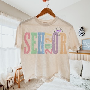 Puede incluir: Una camiseta de manga corta color crema con la palabra SENIOR y el n&uacute;mero 2027 impresos en letras grandes, desgastadas y multicolores. Los colores incluyen rosa, verde, azul, amarillo y morado. Se observa un peque&ntilde;o logotipo circular.