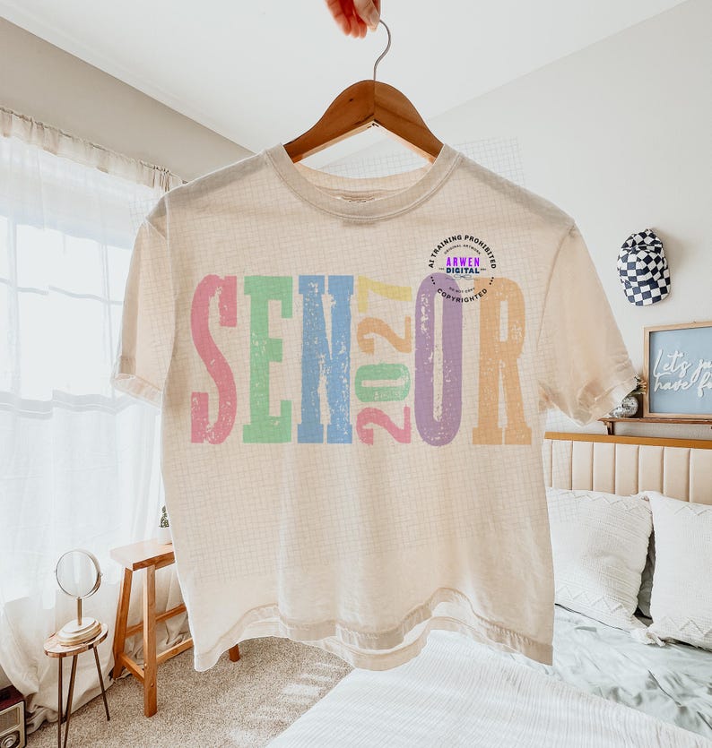 Puede incluir: Una camiseta de manga corta color crema con la palabra SENIOR y el n&uacute;mero 2027 impresos en letras grandes, desgastadas y multicolores. Los colores incluyen rosa, verde, azul, amarillo y morado. Se observa un peque&ntilde;o logotipo circular.
