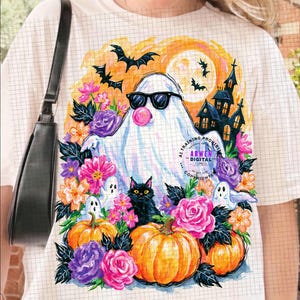 May include: A cream-coloured t-shirt featuring a graphic of a white ghost wearing black sunglasses and blowing a pink bubble. The design includes orange pumpkins, a black cat, purple and pink flowers, bats, and a haunted house silhouette.