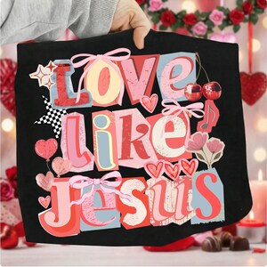 May include: Black tote bag featuring the phrase "Love Like Jesus" in a colourful, collage-style design. The design includes hearts, bows, stars, and cherries. The background is blurred, with Valentine's Day decorations.