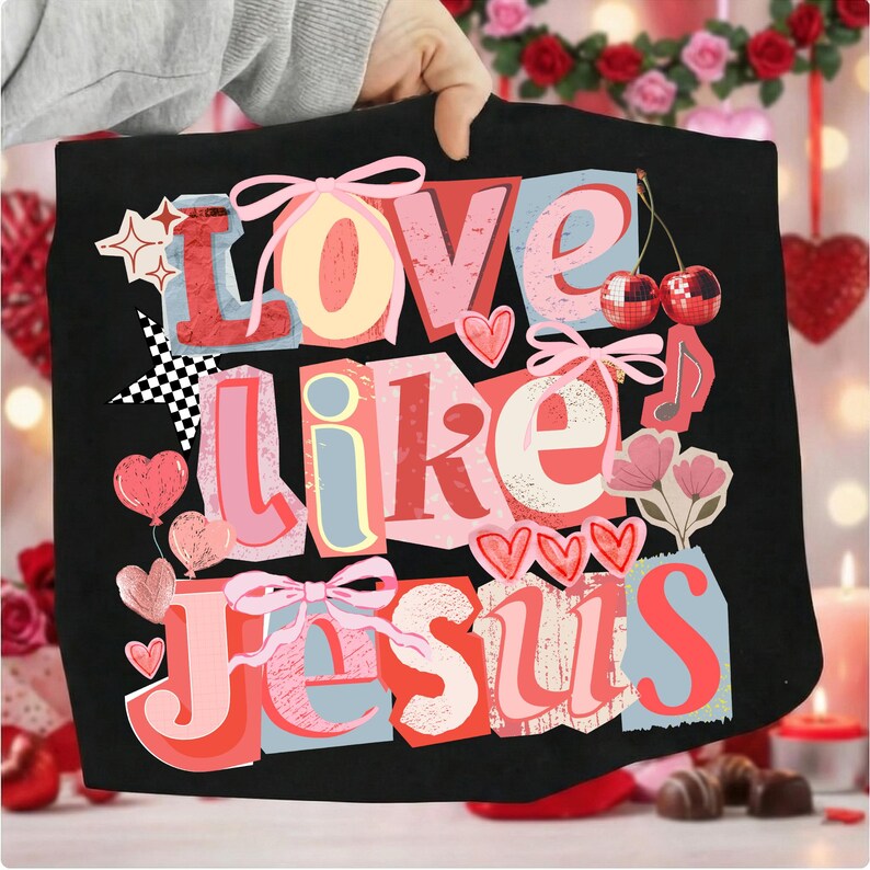 May include: Black tote bag featuring the phrase "Love Like Jesus" in a colourful, collage-style design. The design includes hearts, bows, stars, and cherries. The background is blurred, with Valentine's Day decorations.