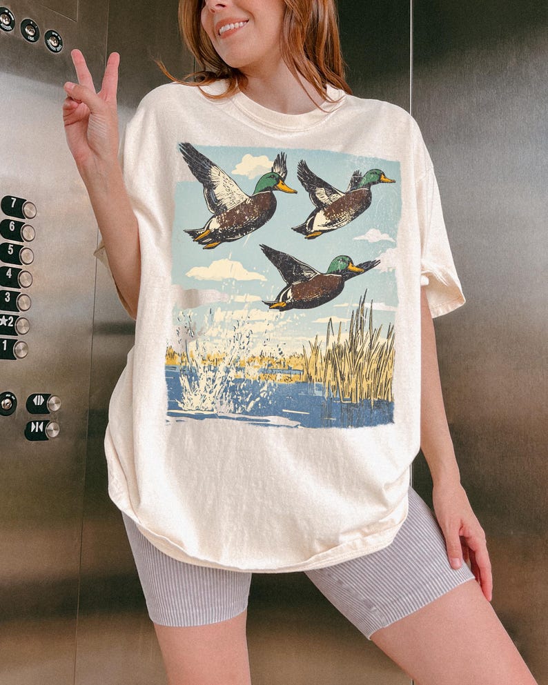 May include: Cream-colored t-shirt featuring a nature scene with three ducks in flight over a body of water. The ducks have brown and green feathers, with a blue sky and yellow reeds in the background. The shirt is oversized.