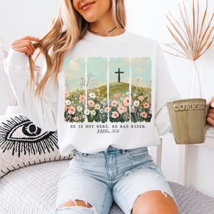 May include: White sweatshirt with a floral design and a cross on a hill. The text "He is not here, He has risen. Matthew 28:6" is printed on the front.