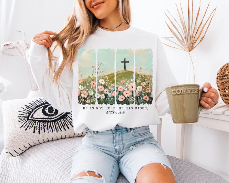 May include: White sweatshirt with a floral design and a cross on a hill. The text "He is not here, He has risen. Matthew 28:6" is printed on the front.