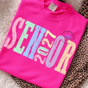 Puede incluir: Una camiseta de manga corta de color rosa brillante con la palabra SENIOR y el n&uacute;mero 2027 en letras de bloque grandes, desgastadas y multicolores con un patr&oacute;n de cuadr&iacute;cula. La camiseta tiene cuello redondo y etiqueta Comfort Colors.