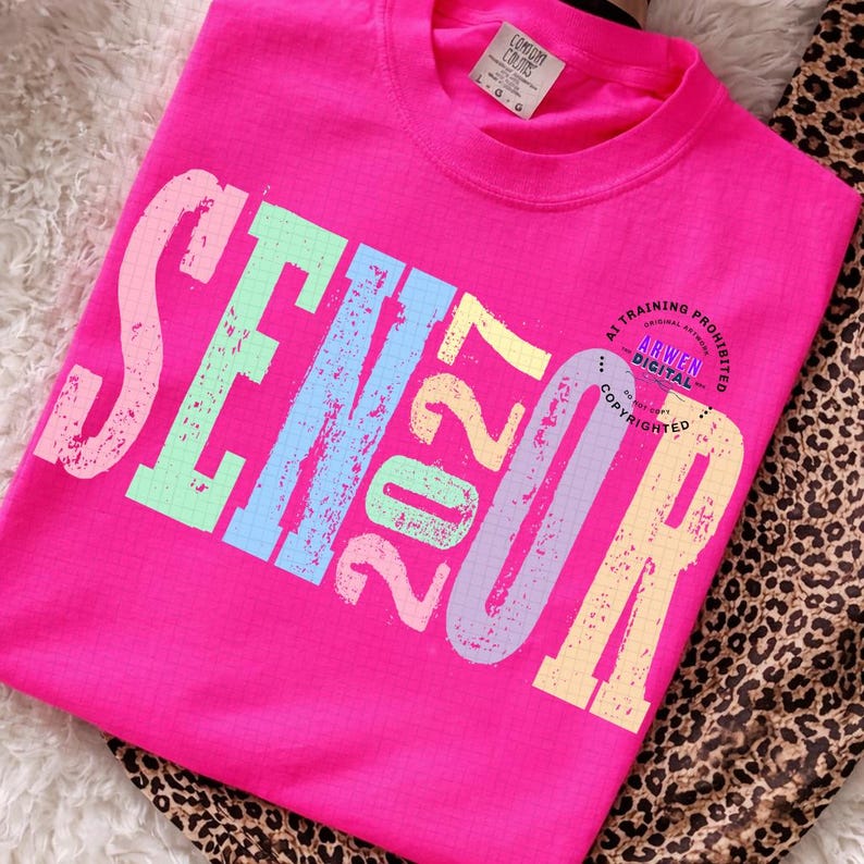 Puede incluir: Una camiseta de manga corta de color rosa brillante con la palabra SENIOR y el n&uacute;mero 2027 en letras de bloque grandes, desgastadas y multicolores con un patr&oacute;n de cuadr&iacute;cula. La camiseta tiene cuello redondo y etiqueta Comfort Colors.