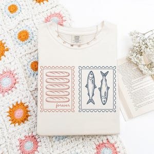 May include: Cream-colored t-shirt featuring two postage stamp designs. One stamp shows six loaves of bread with the word "forever" below. The other stamp displays two fish. A silver necklace and a crocheted blanket are also visible.