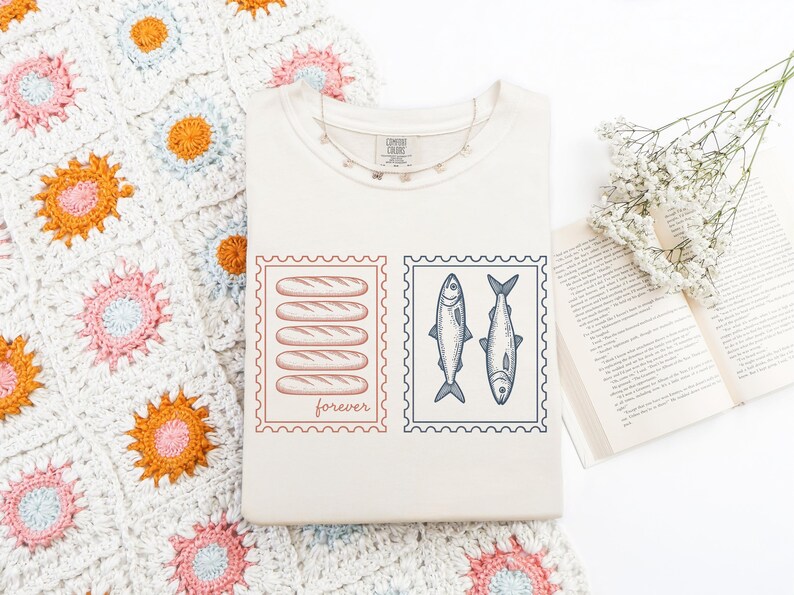 May include: Cream-colored t-shirt featuring two postage stamp designs. One stamp shows six loaves of bread with the word "forever" below. The other stamp displays two fish. A silver necklace and a crocheted blanket are also visible.