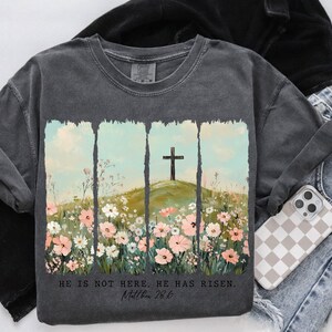 May include: A charcoal grey t-shirt with a floral design and a cross. The text "He is not here. He has risen. Matthew 28:6" is printed on the shirt.