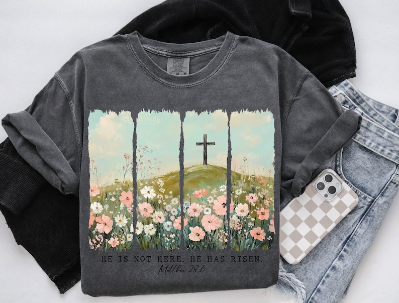 May include: A charcoal grey t-shirt with a floral design and a cross. The text "He is not here. He has risen. Matthew 28:6" is printed on the shirt.