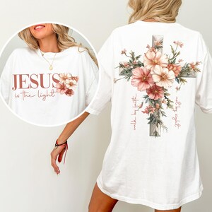 May include: White t-shirt with a floral cross design on the back and the text "Jesus is the light" with floral accents on the front. The text "walk by faith" is written on the cross.