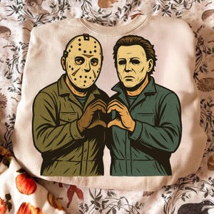 May include: Beige sweatshirt featuring a graphic of two horror movie characters forming a heart shape with their hands. One character wears a hockey mask, the other has dark hair and a stern expression. The figures are in green and brown.