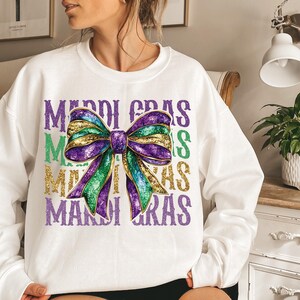 Puede incluir: Sudadera blanca con un lazo de purpurina morado, verde y dorado y el texto "Mardi Gras" repetido tres veces.
