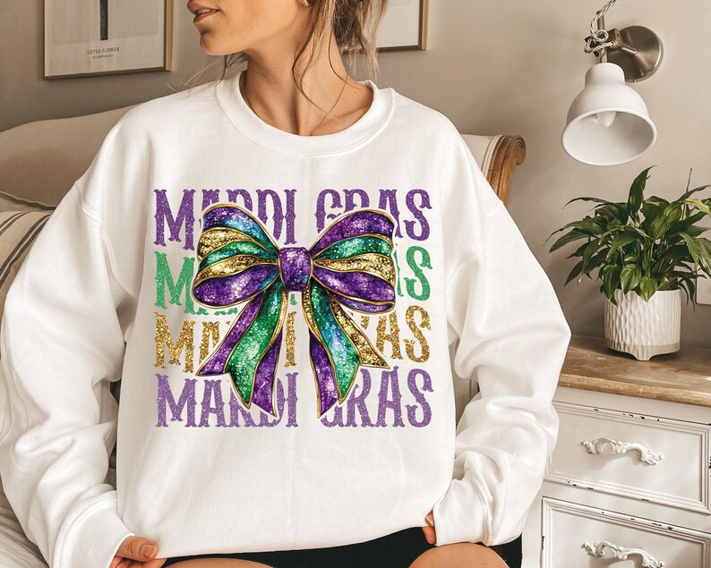 Puede incluir: Sudadera blanca con un lazo de purpurina morado, verde y dorado y el texto "Mardi Gras" repetido tres veces.