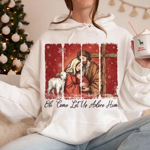 May include: White hoodie featuring a religious illustration with a lamb, a family, and a cross against a red backdrop. The text "Oh Come Let Us Adore Him" is printed below the image. A Christmas tree is in the background.