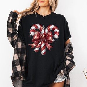 Op de afbeelding: Zwart T-shirt met een kerst suikerstok ontwerp. De afbeelding toont twee rood-wit gestreepte suikerstokken die zijn vastgebonden met een glinsterende rode strik. Het ontwerp heeft een glittereffect, perfect voor de feestdagen.