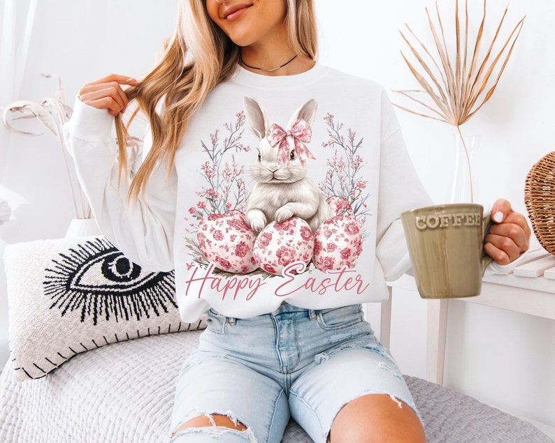 May include: White sweatshirt with a pink and white bunny rabbit holding pink and white floral eggs. The text "Happy Easter" is printed below the bunny.