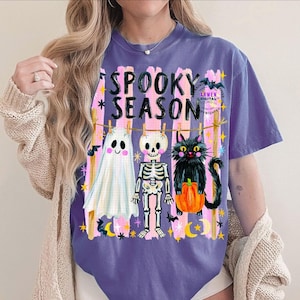 K&ouml;nnte beinhalten: Ein violettes T-Shirt mit dem Text SPOOKY SEASON in schwarzen Buchstaben. Die Grafik zeigt ein wei&szlig;es Gespenst, ein Skelett und eine schwarze Katze mit einem orangefarbenen K&uuml;rbis, die an einer W&auml;scheleine vor rosa vertikalen Streifen h&auml;ngen.