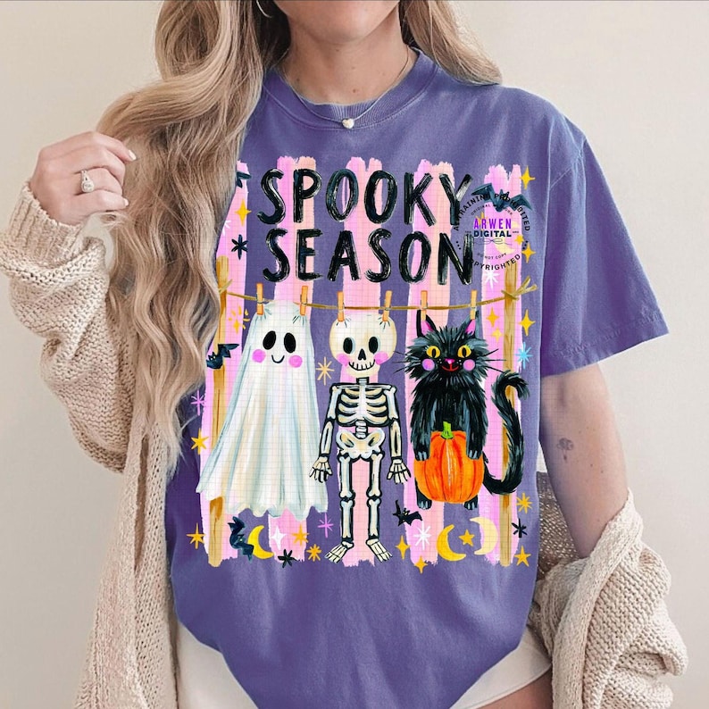 K&ouml;nnte beinhalten: Ein violettes T-Shirt mit dem Text SPOOKY SEASON in schwarzen Buchstaben. Die Grafik zeigt ein wei&szlig;es Gespenst, ein Skelett und eine schwarze Katze mit einem orangefarbenen K&uuml;rbis, die an einer W&auml;scheleine vor rosa vertikalen Streifen h&auml;ngen.