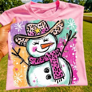 May include: Pink crew neck t-shirt with a snowman design. The snowman has a leopard print cowboy hat and scarf, a carrot nose, and twig arms. The background features colourful brushstrokes and snowflakes.