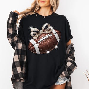 Pode incluir: Camiseta preta com uma bola de futebol marrom e branca com um la&ccedil;o prateado brilhante no topo. A bola de futebol tem costuras brancas e um design prateado brilhante.
