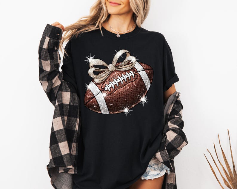 Pode incluir: Camiseta preta com uma bola de futebol marrom e branca com um la&ccedil;o prateado brilhante no topo. A bola de futebol tem costuras brancas e um design prateado brilhante.