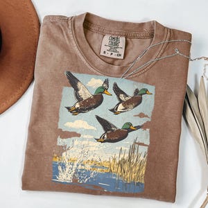 May include: Brown Comfort Colors t-shirt featuring a graphic of three flying ducks over a body of water. The ducks are brown with green heads and yellow beaks. The shirt has a crew neck and a small tag.