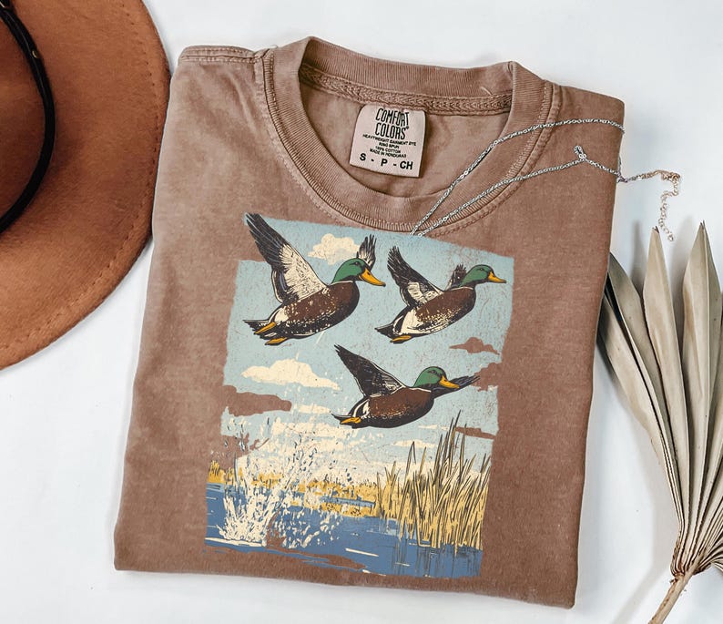 May include: Brown Comfort Colors t-shirt featuring a graphic of three flying ducks over a body of water. The ducks are brown with green heads and yellow beaks. The shirt has a crew neck and a small tag.