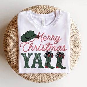 May include: White t-shirt with a festive design. The shirt features the text "Merry Christmas Y'all" in red and green, along with a green cowboy hat and boots. The boots are decorated with candy canes and holly.