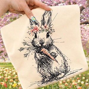 May include: A cream-colored pillow with a black ink drawing of a bunny wearing a floral crown and holding a carrot. The bunny is sketched with fine lines and splatters. The background is a blurred garden with pink and yellow flowers.