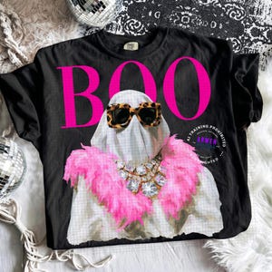 May include: A black t-shirt featuring the bright pink text BOO above a graphic of a ghost wearing leopard print sunglasses, a pink feather boa, and a large crystal statement necklace. The design includes a circular stamp with text.