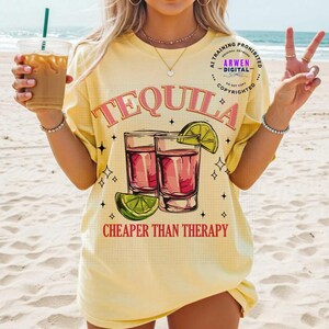 May include: Yellow t-shirt with the word "TEQUILA" in pink script above two shot glasses filled with a red liquid and a lime wedge. The text "CHEAPER THAN THERAPY" is below the glasses. The shirt has a relaxed fit.