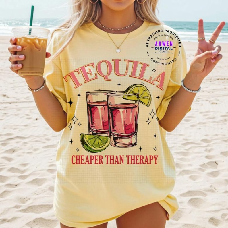 May include: Yellow t-shirt with the word "TEQUILA" in pink script above two shot glasses filled with a red liquid and a lime wedge. The text "CHEAPER THAN THERAPY" is below the glasses. The shirt has a relaxed fit.
