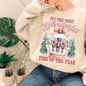 May include: A beige sweatshirt with a Christmas-themed design. The design features a snow globe with three nutcracker figurines inside. The text on the sweatshirt reads "It's the most Wonderful Time of the Year."