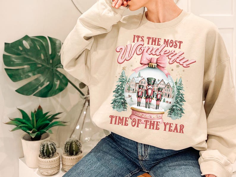 May include: A beige sweatshirt with a Christmas-themed design. The design features a snow globe with three nutcracker figurines inside. The text on the sweatshirt reads "It's the most Wonderful Time of the Year."